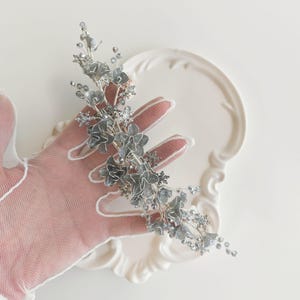 Op de afbeelding: Een zilveren bloemen haaraccessoire met gelaagde bloemblaadjes en sprankelende accenten. Het stuk heeft een flexibele draadbasis en kleine, met kralen bezette versieringen, geschikt voor bruids- of formele gelegenheden.