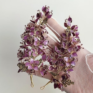 Puede incluir: Un conjunto de cinco pinzas para el pelo con forma de flor morada con detalles de alambre dorado. Las pinzas están hechas de alambre y tienen un aspecto delicado y femenino.