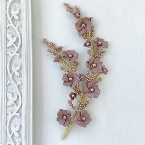 Puede incluir: Un accesorio decorativo para el cabello con flores de color malva y cuentas centrales, acentuado por ramas de alambre en tono dorado y pequeñas cuentas malvas a juego. El arreglo floral tiene una estructura vertical de enredadera.