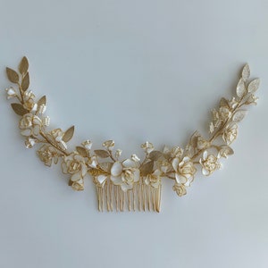 Puede incluir: Peine de pelo dorado con flores blancas y detalles de perlas. El peine está decorado con un diseño floral y tiene un aspecto delicado y femenino.