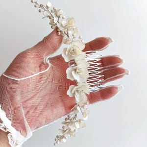 Puede incluir: Peine de pelo floral blanco con detalles de perlas. El peine tiene una base plateada y está decorado con flores blancas y perlas.