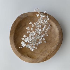 Puede incluir: Un delicado accesorio floral plateado para el cabello con pequeñas flores blancas y detalles de perlas. El adorno para el cabello se presenta en un cuenco de madera con un acabado marrón claro. El diseño floral es intrincado y detallado.
