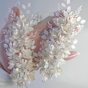 Puede incluir: Dos pinzas para el pelo florales blancas con detalles de perlas y tallos de alambre plateado. Las pinzas están hechas de delicadas flores blancas con un acabado nacarado.