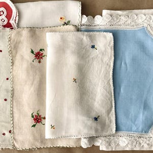 May include: A collection of vintage-style fabric napkins and doilies featuring various designs, including red embroidered dots, floral cross-stitch patterns, a solid light blue centre with white lace trim, and geometric red and blue border stitching.