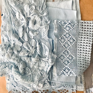 May include: Assortment of light blue lace and fabric swatches. The collection includes floral embroidered lace, crocheted lace, and plain fabric pieces. The textiles vary in texture and design, showcasing intricate details and patterns. Ideal for craft projects.