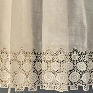 Puede incluir: Primer plano de cortinas de encaje blanco con borde festoneado. El borde inferior presenta un patr&oacute;n repetitivo de dise&ntilde;os en espiral dentro de un marco geom&eacute;trico. La tela parece semitransparente.
