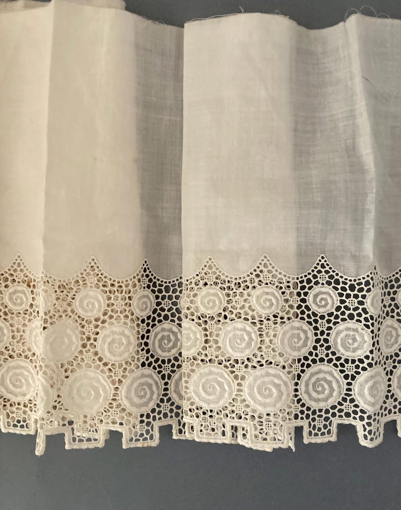 Puede incluir: Primer plano de cortinas de encaje blanco con borde festoneado. El borde inferior presenta un patr&oacute;n repetitivo de dise&ntilde;os en espiral dentro de un marco geom&eacute;trico. La tela parece semitransparente.
