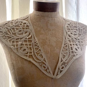 May include: Cream-colored lace collar with an intricate, openwork design. The collar is displayed on a vintage dress form, showcasing its delicate patterns and textures. The lace features swirling and geometric motifs, creating an elegant, vintage aesthetic.