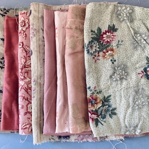 May include: A collection of fabric swatches featuring various textures and patterns, including solid dusty rose, floral prints in red and cream, a beige linen-like weave, and a cream-coloured textile with a detailed multi-coloured floral bouquet.