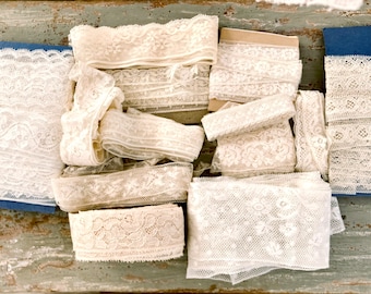 May include: A collection of ivory-colored lace ribbons in various widths and patterns. The lace is rolled and stacked, showcasing floral and geometric designs. Some rolls are on cardboard spools, while others are loose. The background is a distressed, weathered wooden surface.