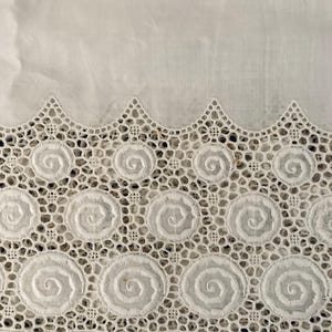 Puede incluir: Primer plano de tela blanca con un borde de encaje calado detallado. El encaje presenta un patr&oacute;n repetitivo de dise&ntilde;os en espiral dentro de marcos circulares, creando un borde delicado y texturizado. La parte superior de la imagen muestra tela blanca lisa.