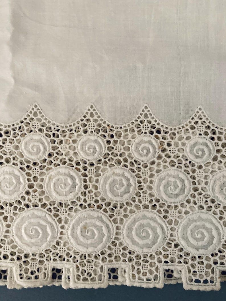 Puede incluir: Primer plano de tela blanca con un borde de encaje calado detallado. El encaje presenta un patr&oacute;n repetitivo de dise&ntilde;os en espiral dentro de marcos circulares, creando un borde delicado y texturizado. La parte superior de la imagen muestra tela blanca lisa.