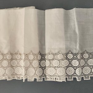 Puede incluir: Tela blanca con un borde de encaje decorativo con un dise&ntilde;o de espiral repetitivo. El encaje est&aacute; intrincadamente detallado, agregando un toque delicado a la tela. La tela est&aacute; doblada, mostrando la textura y el patr&oacute;n del encaje.