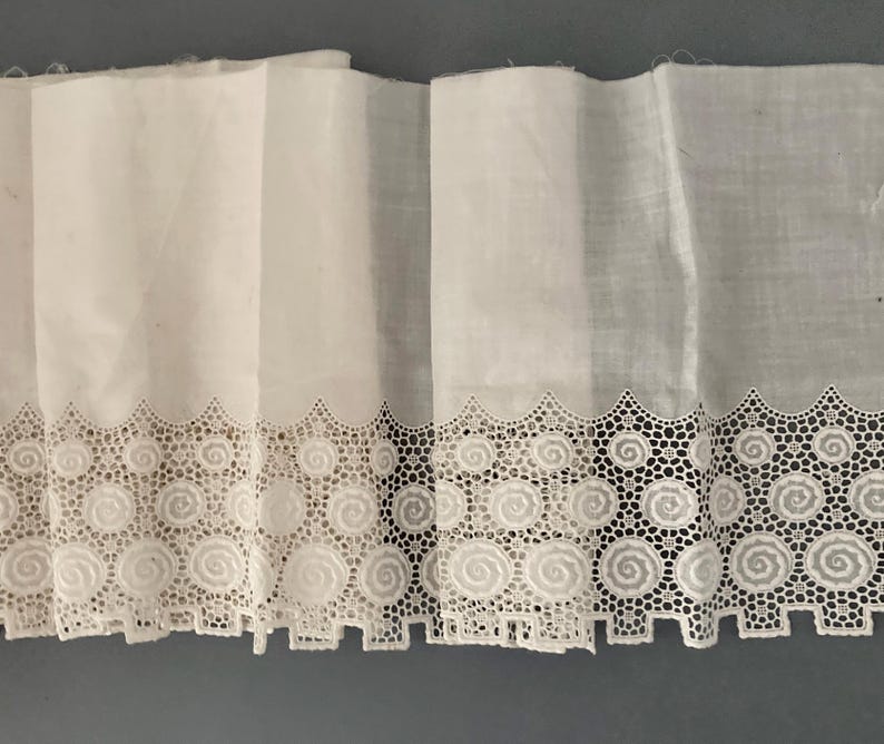 Puede incluir: Tela blanca con un borde de encaje decorativo con un dise&ntilde;o de espiral repetitivo. El encaje est&aacute; intrincadamente detallado, agregando un toque delicado a la tela. La tela est&aacute; doblada, mostrando la textura y el patr&oacute;n del encaje.