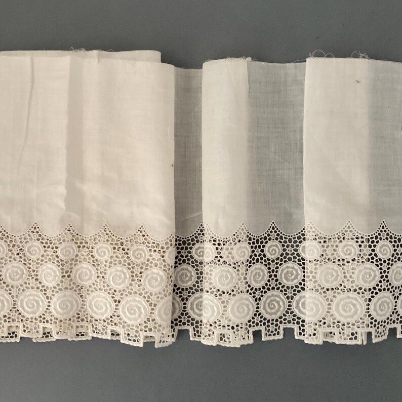Puede incluir: Tela blanca con un borde de encaje calado decorativo con un dise&ntilde;o de espiral repetitivo. La tela est&aacute; doblada, mostrando m&uacute;ltiples capas y una visi&oacute;n de la textura. El encaje tiene un borde festoneado.