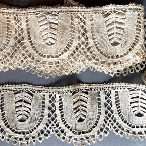 Puede incluir: Dos tiras de encaje beige antiguo con un borde festoneado. El encaje presenta un patrón repetitivo de diseños arqueados con un motivo central en forma de hoja. La delicada construcción calada es visible en todo el encaje.