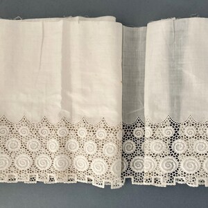 Puede incluir: Tela blanca con un borde de encaje calado decorativo con un dise&ntilde;o en espiral. La tela est&aacute; doblada, mostrando la textura y los detalles del encaje. La est&eacute;tica general es vintage o antigua.