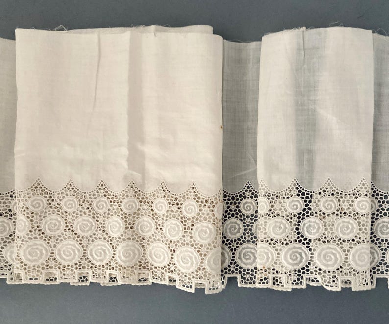 Puede incluir: Tela blanca con un borde de encaje calado decorativo con un dise&ntilde;o en espiral. La tela est&aacute; doblada, mostrando la textura y los detalles del encaje. La est&eacute;tica general es vintage o antigua.