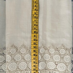 Puede incluir: Tela blanca con un borde de encaje decorativo con un dise&ntilde;o en espiral. Una cinta m&eacute;trica amarilla muestra que la tela mide aproximadamente 61 cm de largo. La tela probablemente se utiliza para coser o hacer manualidades.