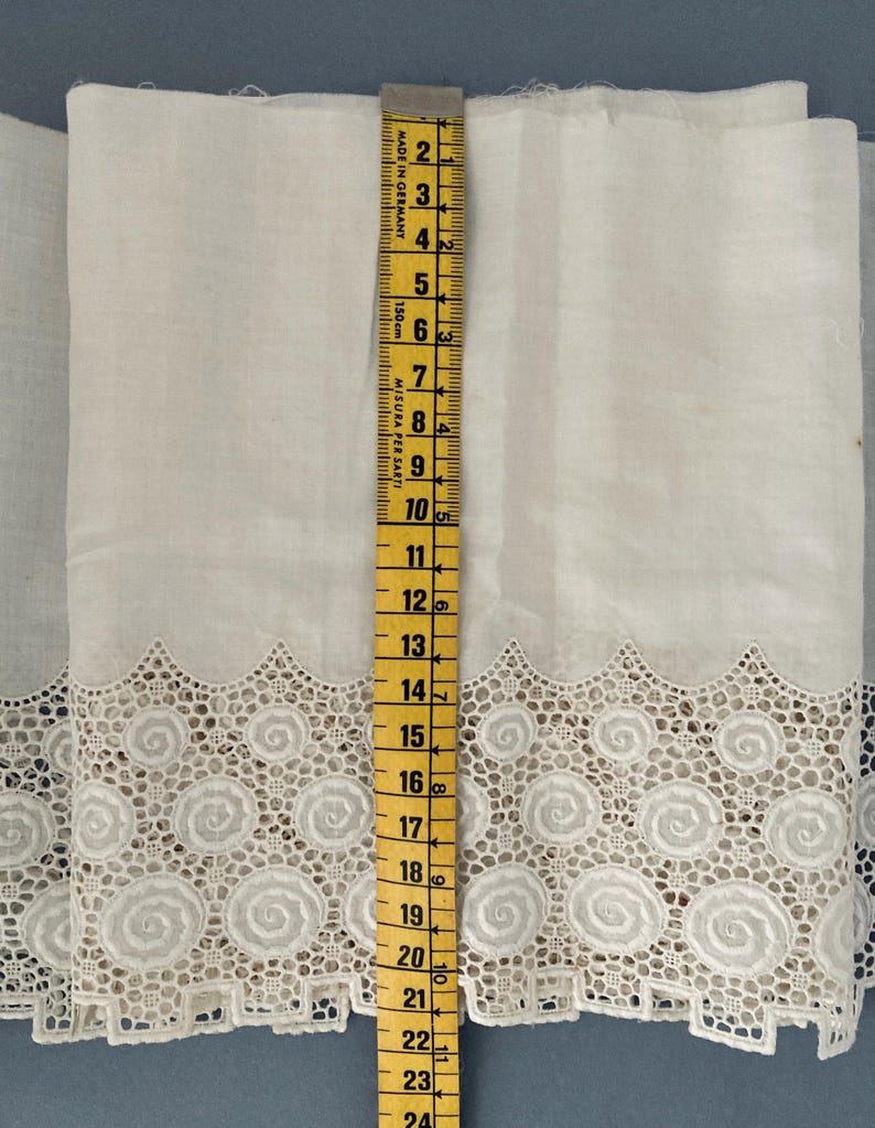 Puede incluir: Tela blanca con un borde de encaje decorativo con un dise&ntilde;o en espiral. Una cinta m&eacute;trica amarilla muestra que la tela mide aproximadamente 61 cm de largo. La tela probablemente se utiliza para coser o hacer manualidades.