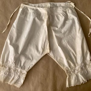 以下が含まれることがあります： 股割れデザインの白いコットン製ドロワーズ。ウエストには調節可能なサイドタイがあり、脚の開口部は複雑なアイレットレースのトリムと装飾的なジグザグ模様で仕上げられています。ゆったりとしたギャザーのシルエットが特徴です。
