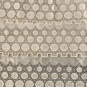 Puede incluir: Primer plano de tela de encaje blanco con un dise&ntilde;o de espiral repetitivo y bordes festoneados. El encaje est&aacute; detallado con peque&ntilde;os agujeros, creando una textura delicada. La tela parece ser vintage o antigua.