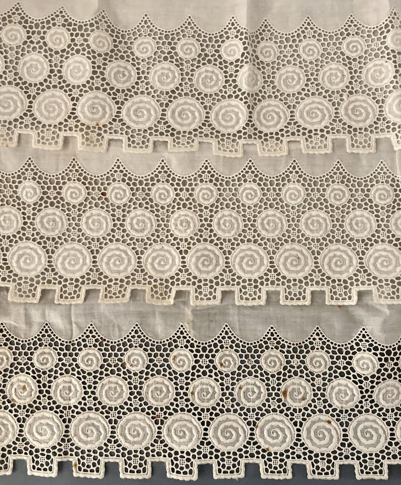 Puede incluir: Primer plano de tela de encaje blanco con un dise&ntilde;o de espiral repetitivo y bordes festoneados. El encaje est&aacute; detallado con peque&ntilde;os agujeros, creando una textura delicada. La tela parece ser vintage o antigua.