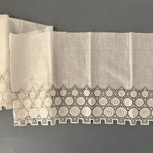 Puede incluir: Dos paneles de tela blanca con un borde calado decorativo con un dise&ntilde;o repetitivo de espirales y cuadrados. La tela est&aacute; doblada y parece ser un material ligero, posiblemente lino o algod&oacute;n.