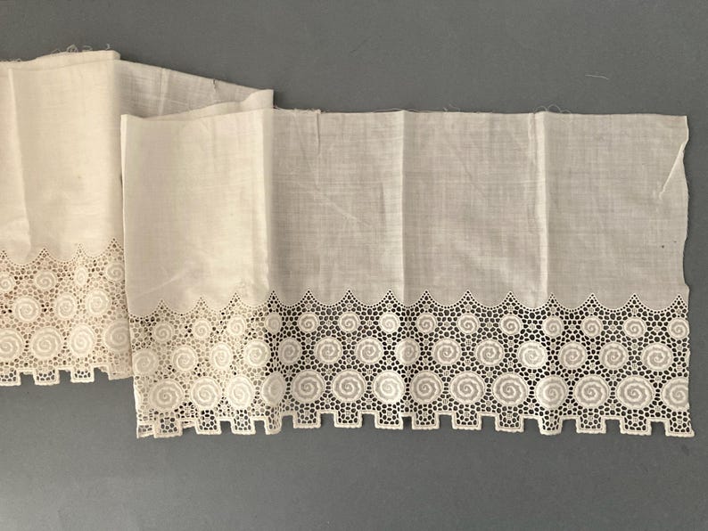 Puede incluir: Dos paneles de tela blanca con un borde calado decorativo con un dise&ntilde;o repetitivo de espirales y cuadrados. La tela est&aacute; doblada y parece ser un material ligero, posiblemente lino o algod&oacute;n.
