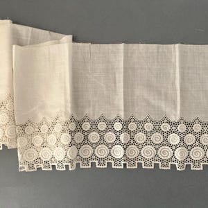 Puede incluir: Tela blanquecina con un borde decorativo calado. El borde presenta un patr&oacute;n repetitivo de espirales circulares y formas geom&eacute;tricas. La tela est&aacute; doblada, mostrando la textura y los detalles del dise&ntilde;o. La est&eacute;tica general es vintage o antigua.