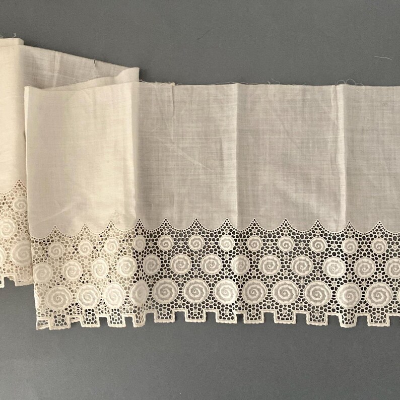 Puede incluir: Tela blanquecina con un borde decorativo calado. El borde presenta un patr&oacute;n repetitivo de espirales circulares y formas geom&eacute;tricas. La tela est&aacute; doblada, mostrando la textura y los detalles del dise&ntilde;o. La est&eacute;tica general es vintage o antigua.