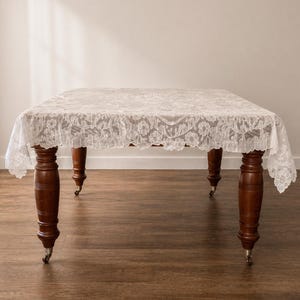 May include: A wooden table with a white lace tablecloth. The table has dark brown, turned legs with small caster wheels. The lace tablecloth has a floral pattern. The table is approximately 120 cm long.