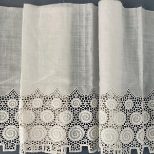 Puede incluir: Tela blanca con un borde de encaje decorativo. El encaje presenta un patr&oacute;n repetitivo de dise&ntilde;os en espiral y bordes festoneados. La tela parece ligera y semitransparente, adecuada para cortinas u otros proyectos de decoraci&oacute;n del hogar.