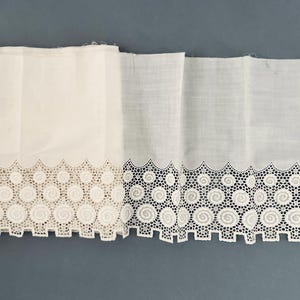 Puede incluir: Dos paneles de tela blanca con un borde de encaje decorativo. El encaje presenta un patr&oacute;n repetitivo de espirales circulares y un borde festoneado. La tela parece ser de algod&oacute;n o lino, con una superficie ligeramente texturizada.