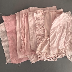May include: A selection of pink fabric samples with diverse textures and designs. The swatches feature lace, embroidered elements, and various pink hues, evoking a vintage or romantic feel. The fabrics are displayed on a grey surface.