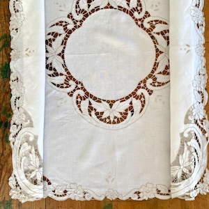 Könnte beinhalten: Ein weißer, rechteckiger Tischläufer mit aufwendiger Lochstickerei und Spitzendetails. Das Design umfasst ein zentrales kreisförmiges Motiv, das von Blumenmustern und gebogten Kanten mit durchbrochenen Stichen im gesamten Stoff umgeben ist.