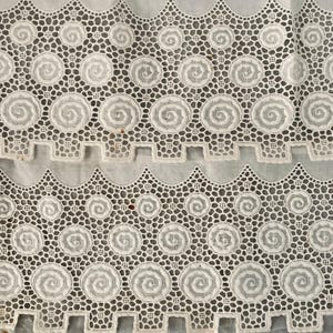 Puede incluir: Primer plano de tela de encaje blanco con un patr&oacute;n repetitivo de dise&ntilde;os en espiral y bordes festoneados. El encaje tiene una textura delicada y calada, creando una apariencia detallada e intrincada. La tela se utiliza probablemente para ropa o decoraci&oacute;n del hogar.