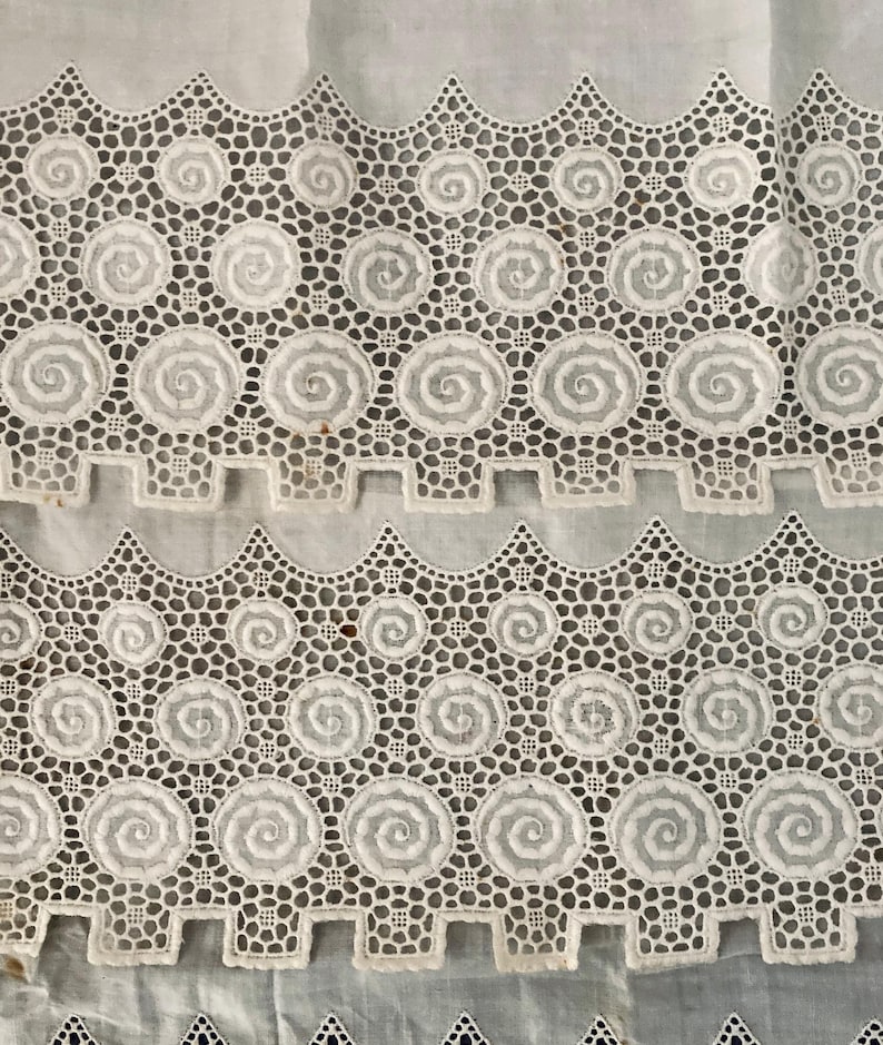 Puede incluir: Primer plano de tela de encaje blanco con un patr&oacute;n repetitivo de dise&ntilde;os en espiral y bordes festoneados. El encaje tiene una textura delicada y calada, creando una apariencia detallada e intrincada. La tela se utiliza probablemente para ropa o decoraci&oacute;n del hogar.