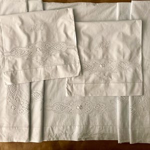 May include: White bed linens set, including a flat sheet and two pillowcases. The pillowcases and the top of the flat sheet feature embroidered floral and geometric lace details. The pillowcases are monogrammed with the letters "MC".