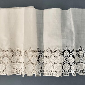 Puede incluir: Tela blanca con un ribete de encaje decorativo con un dise&ntilde;o de espiral repetido. El ribete de encaje est&aacute; festoneado a lo largo del borde inferior. La tela est&aacute; doblada, creando l&iacute;neas verticales. El fondo es azul liso y apagado.