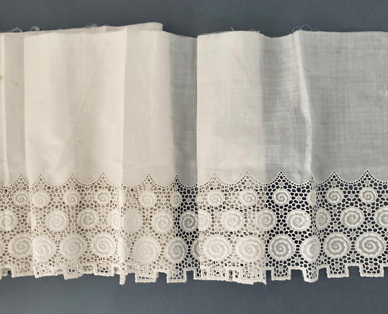 Puede incluir: Tela blanca con un ribete de encaje decorativo con un dise&ntilde;o de espiral repetido. El ribete de encaje est&aacute; festoneado a lo largo del borde inferior. La tela est&aacute; doblada, creando l&iacute;neas verticales. El fondo es azul liso y apagado.