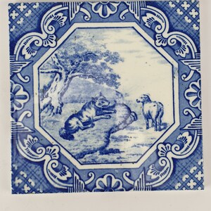 May include: A blue and white ceramic tile with a decorative border and a scene of a dog and a sheep in a field. The tile is in the style of Delftware.