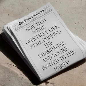 May include: A stack of newspapers with the headline "NOW THAT WE'RE OFFICIALLY LIVE, WE'RE POPPING THE CHAMPAGNE AND YOU'RE INVITED TO THE PARTY!" The newspaper is titled "The Business Times" and is a special edition.