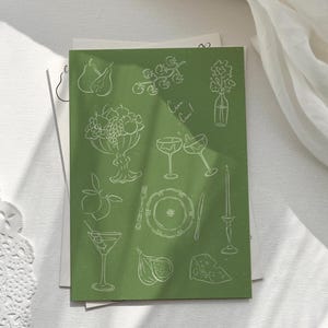 May include: A green card with white line drawings of various objects, including fruit, glasses, a plate, and a candle. The text "Cheers!" is written on the card.