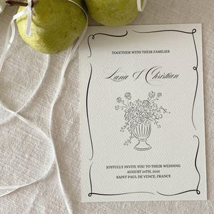 May include: A white wedding invitation with black calligraphy announcing the wedding of Lama and Christian. The invitation includes the date, August 24th, 2025, and the location, Saint Paul de Vence, France.