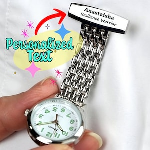 Op de afbeelding: Een zilveren horloge met een witte wijzerplaat en zwarte cijfers. Het horloge heeft een gepersonaliseerd opschrift met de tekst "Anastaisha Resilience Warrior".