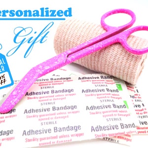 Könnte beinhalten: Eine rosa medizinische Schere mit einer Rolle rosa Mullbinde und einem Stapel Pflaster, auf denen "Adhesive Bandage" und "Sterility guaranteed unless wrapper opened or damaged STERILE" steht.