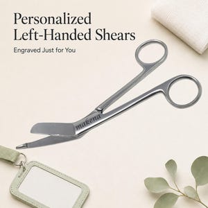 May include: Silver-colored metal trauma shears with a bent blade design and the name makena engraved on the handle. The text Personalized Left-Handed Shears Engraved Just for You is visible above the shears, which are positioned near a badge holder.