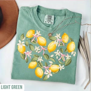 May include: Light green t-shirt with a heart-shaped lemon design. The design includes yellow lemons, white flowers, pink ribbons, and green leaves. The text "Fuck It" is printed on each lemon. A silver necklace is also visible.