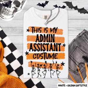 May include: A white t-shirt featuring the black text "THIS IS MY ADMIN ASSISTANT COSTUME" layered over four horizontal orange brushstroke stripes. Below the text are five black skeleton illustrations, each holding office supplies, accented by small black stars.